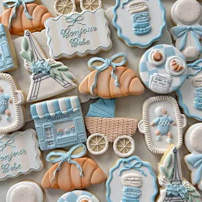 Parisian-themed baby shower cookie set with a "Bonjour Bebe" plaque, Eiffel Tower, baby carriage, croissants, macarons, and a patisserie storefront in soft blue and white.