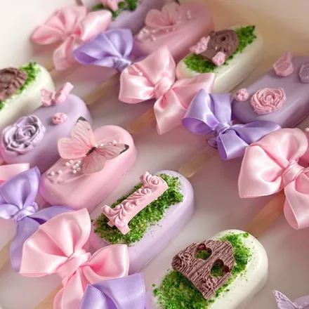 A dozen pastel cakesicles in purple, pink, and white, decorated with satin bows, butterflies, roses, and a number-one topper.
