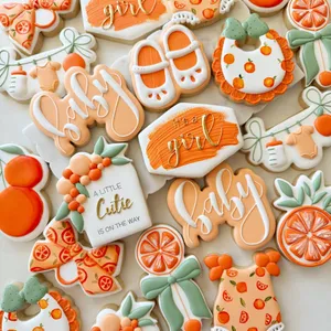 Orange citrus-themed "It's a Girl" baby shower cookie set with orange slices, a baby onesie, tiny shoes, a bib, and "A Little Cutie" lettering in peach and sage green.