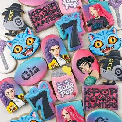 K-pop themed birthday cookie set for "Gia" with animated idol characters, a soda can, cat mascots, and "K-Pop Demon Hunters" lettering in pink, purple, and teal.