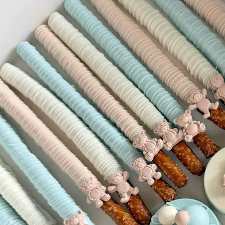 Chocolate-covered pretzel rods in pastel blue, cream, and pink, each topped with a small teddy bear for a baby shower.