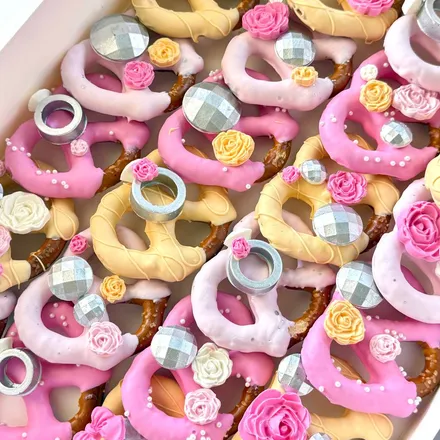 Chocolate-covered pretzel twists in pink, yellow, and white, decorated with sugar roses and faceted "engagement ring" toppers.