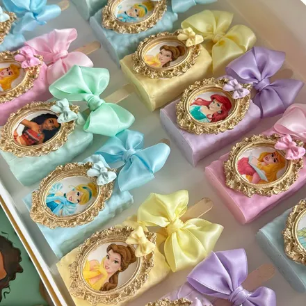 Rice krispie treat pops dipped in pastel colors, each topped with a gold-framed Disney princess portrait.