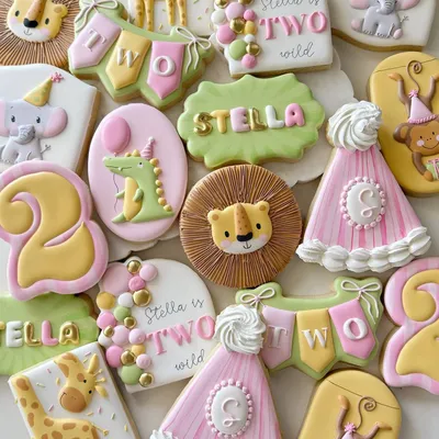 Safari-themed 2nd birthday cookie set for "Stella" with a lion, giraffe, elephant, monkeys, party hats, and "Stella is TWO Wild" lettering in pink, green, and gold.